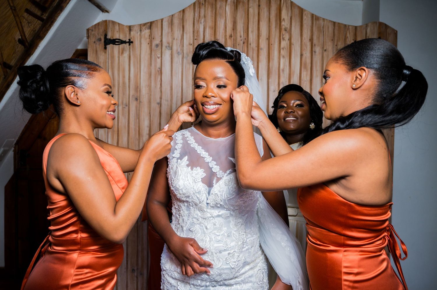 Bridesmaids in orange satin dresses help a bride in a lace wedding gown with her accessories.