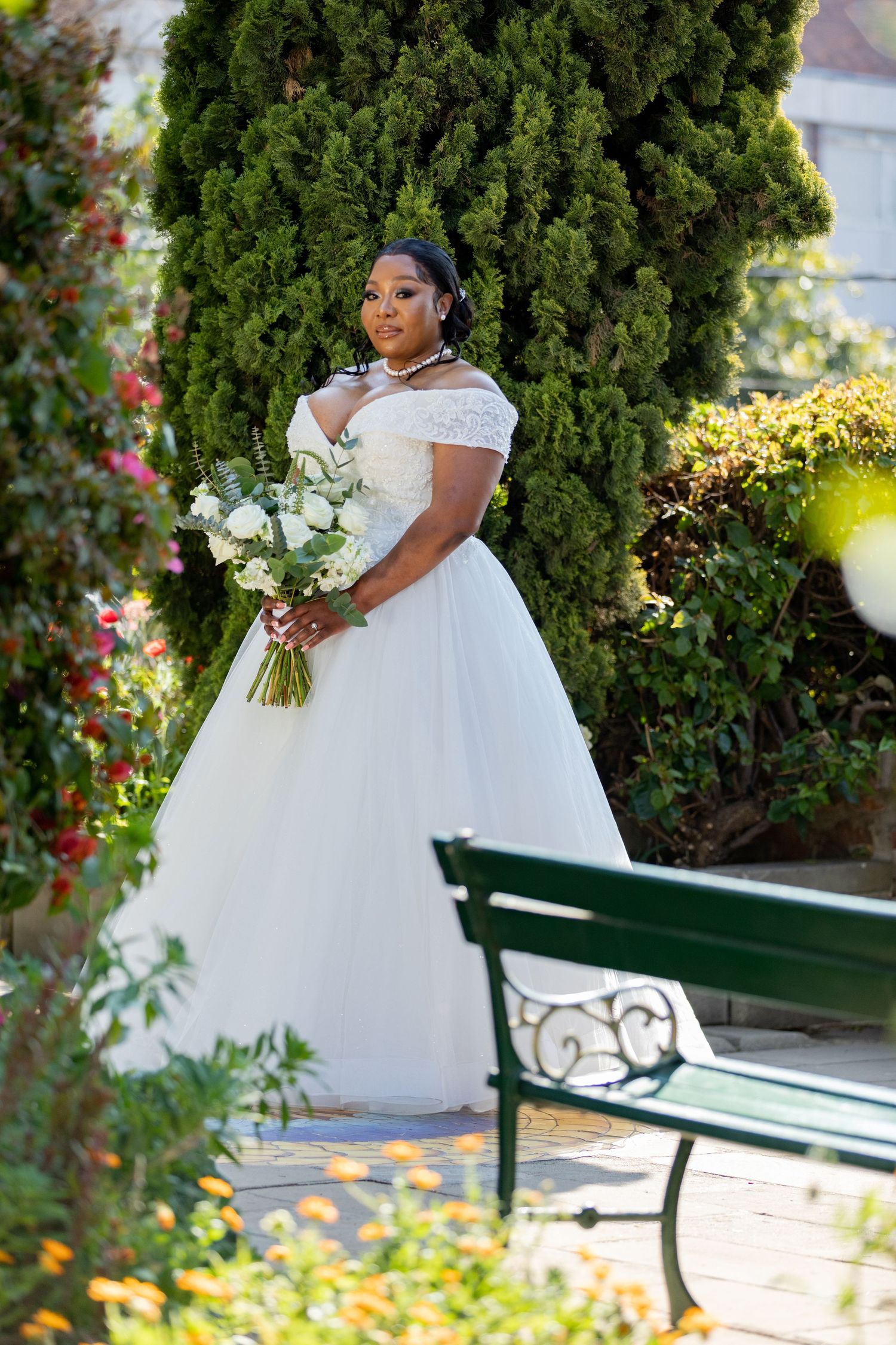 Bride in white off-shoulder ball gown holding white bouquet standing near evergreen tree outdoors.