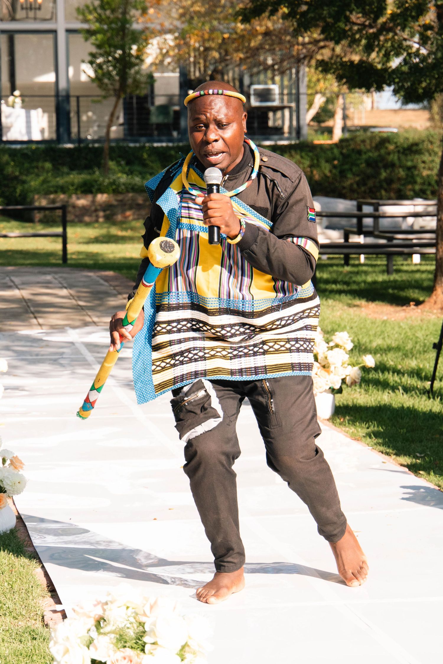 Barefoot performer in traditional African attire speaking into a microphone at an outdoor wedding ceremony.