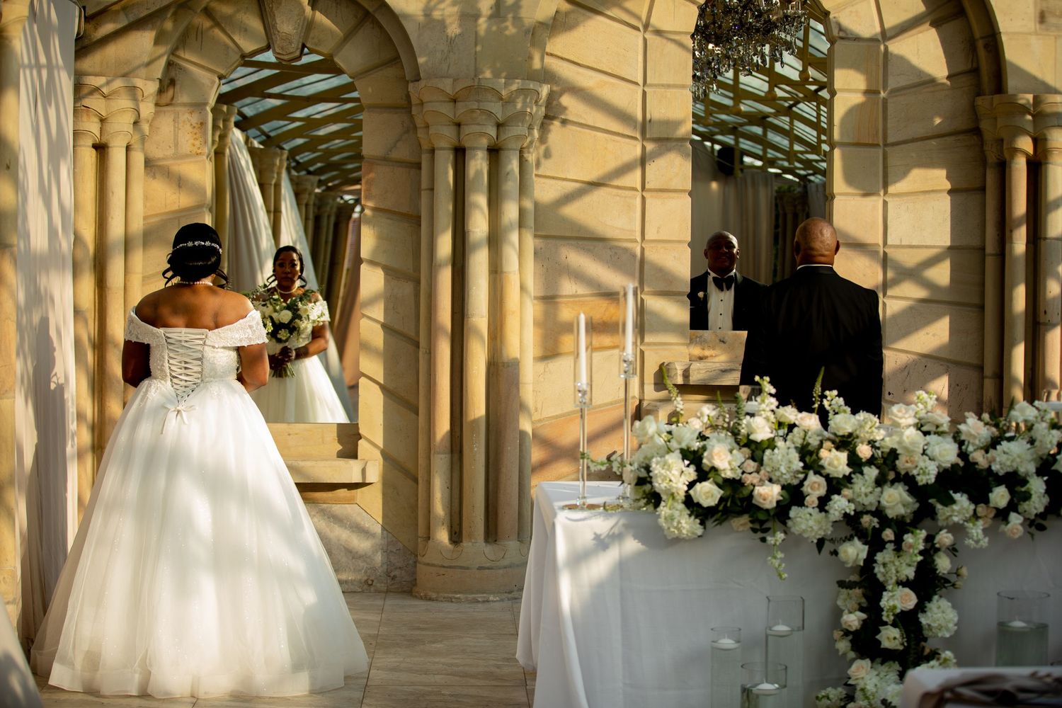 Bride in white ball gown walks toward altar adorned with white floral arrangements at outdoor ceremony.