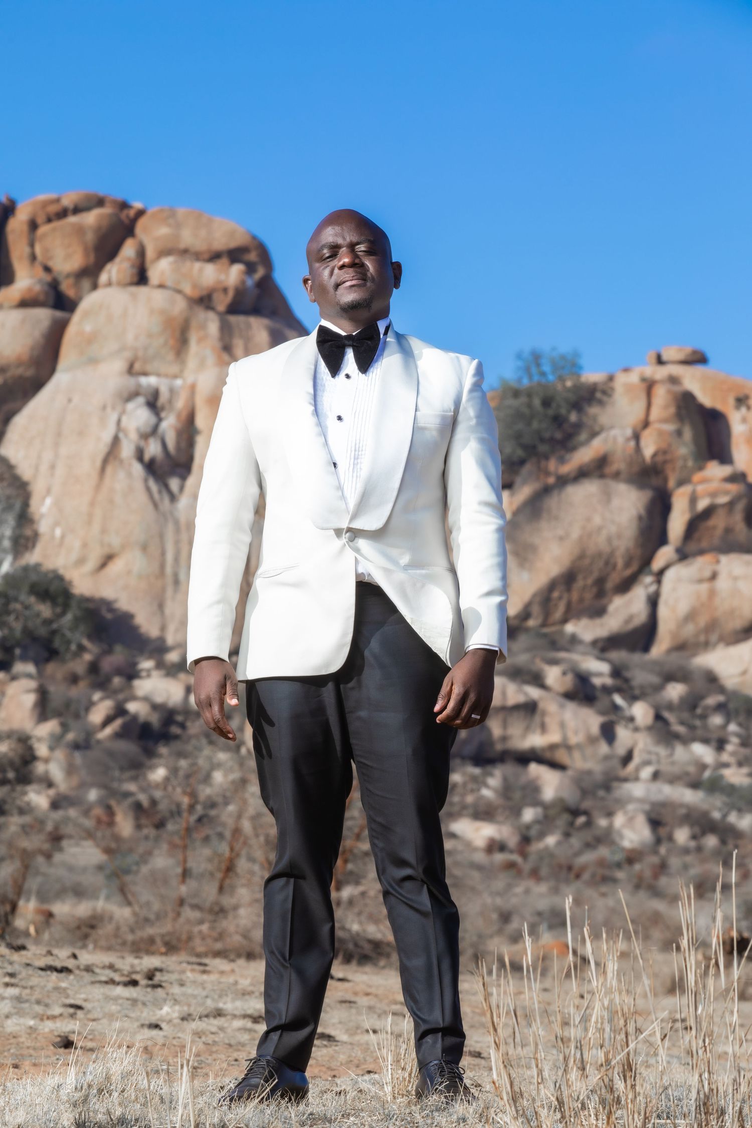 African groom in white tuxedo with bow tie standing in rocky desert landscape.