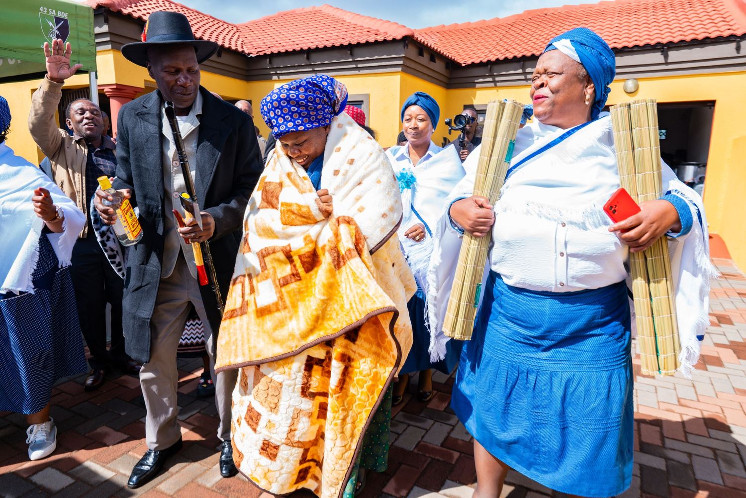 A woman wrapped in a colorful blanket being escorted by elders during a traditional African ceremony.