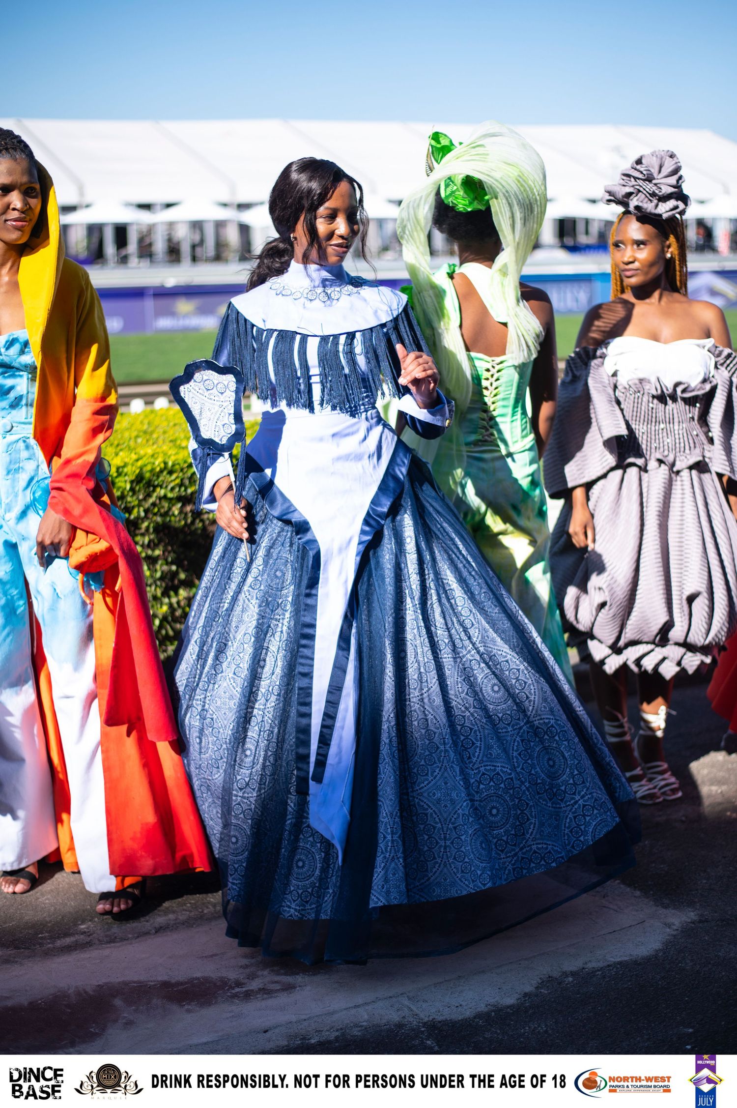Model showcases stunning blue Victorian-style ball gown at an outdoor fashion event at a racecourse.