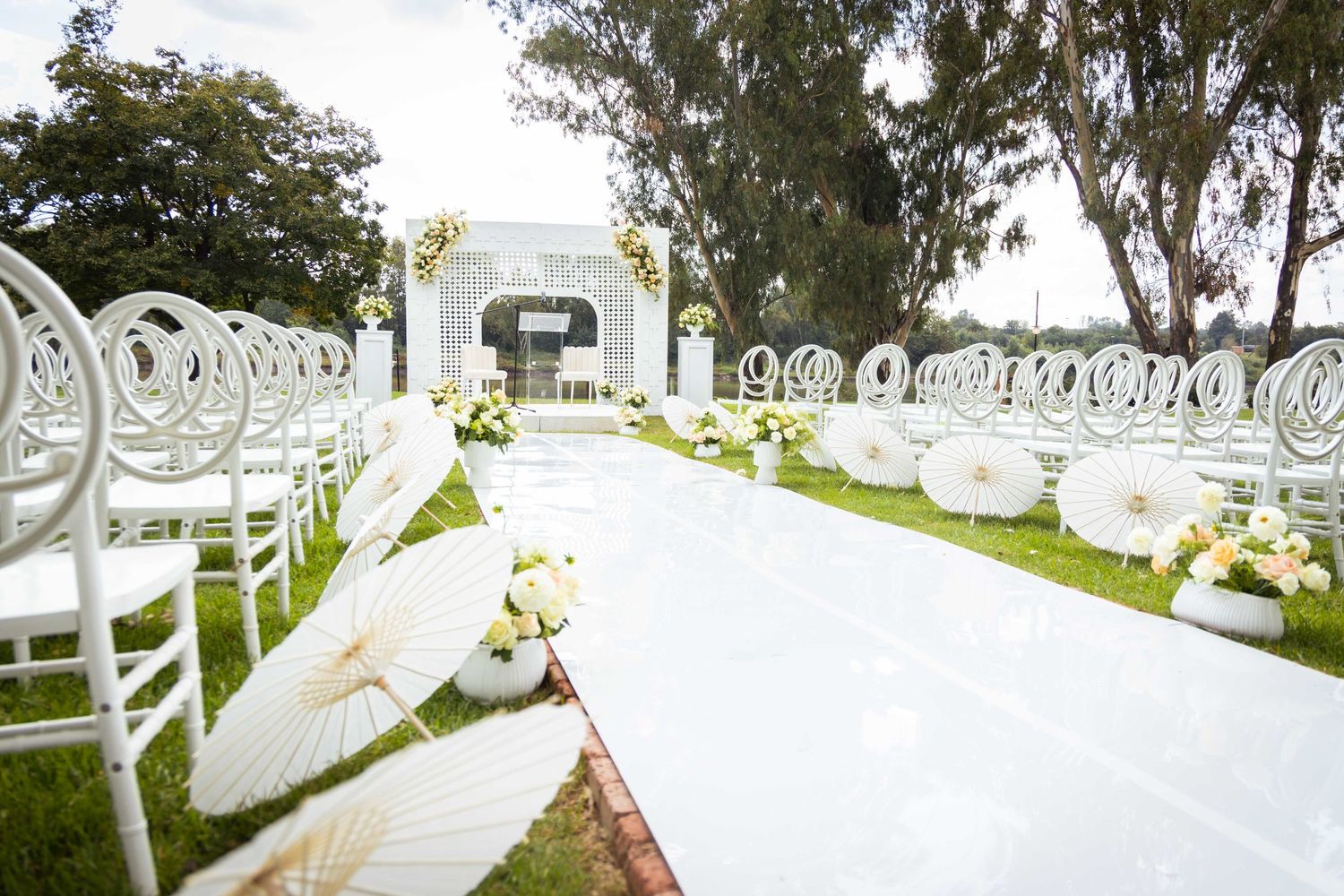 Elegant outdoor wedding ceremony setup with white chairs, parasols, and floral aisle.