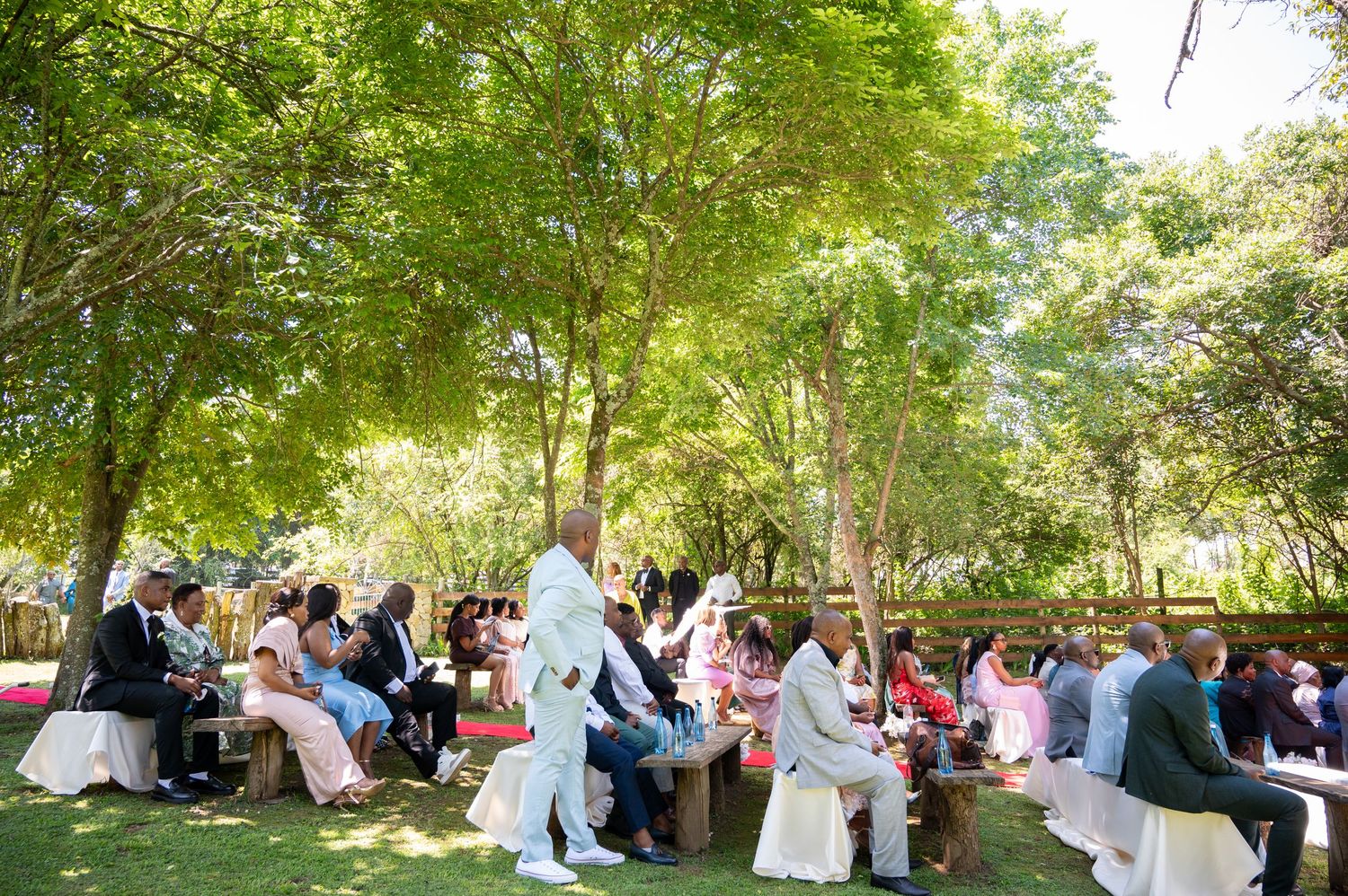 Guests seated outdoors under lush green trees during a wedding ceremony.
