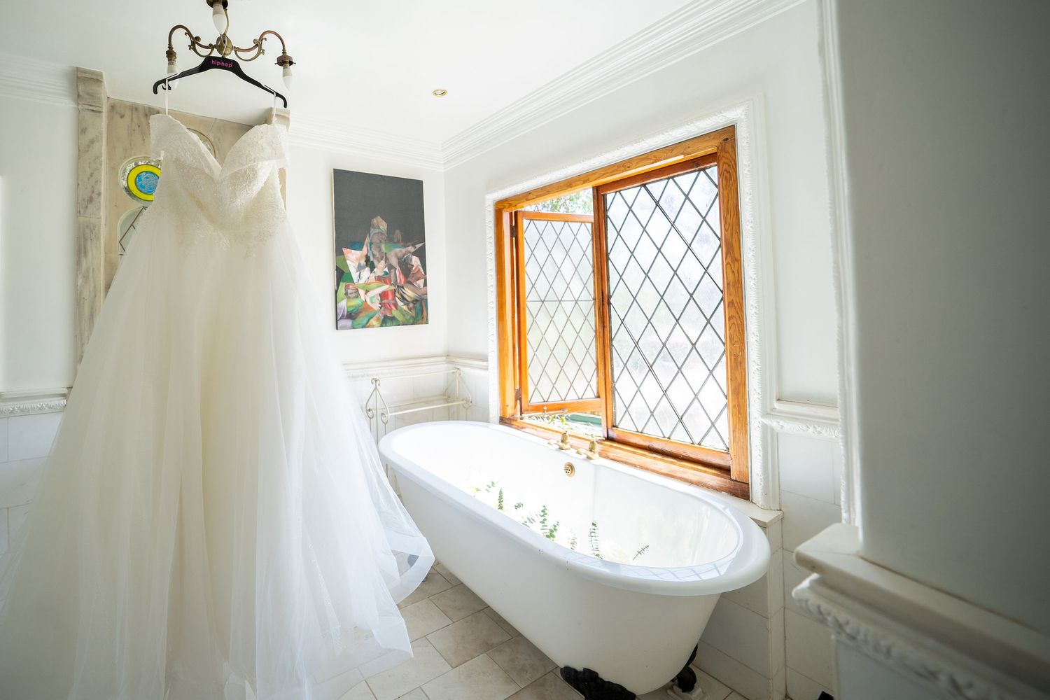 Elegant strapless wedding gown hanging in luxurious white bathroom with clawfoot tub and diamond-paned window.