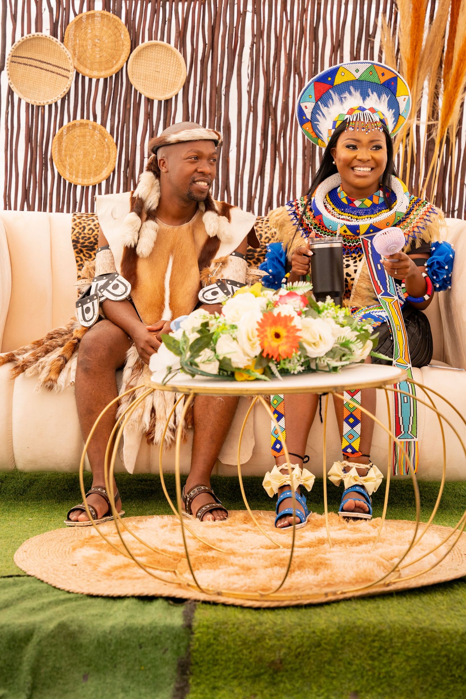 Zulu couple in traditional beaded ceremonial attire sit together on sofa with flowers during cultural wedding ceremony.