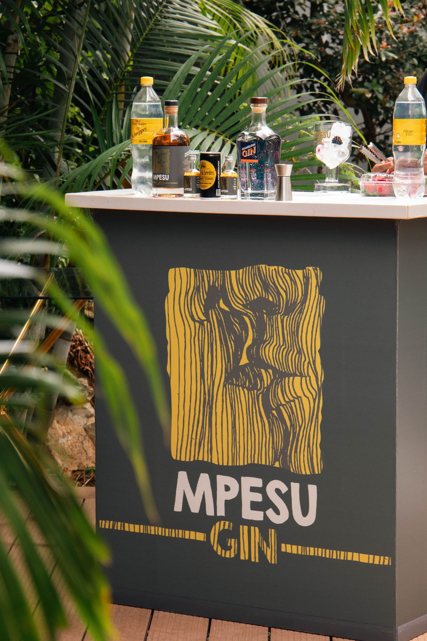 Mpesu Gin outdoor bar setup surrounded by tropical plants with bottles displayed on a grey branded counter.