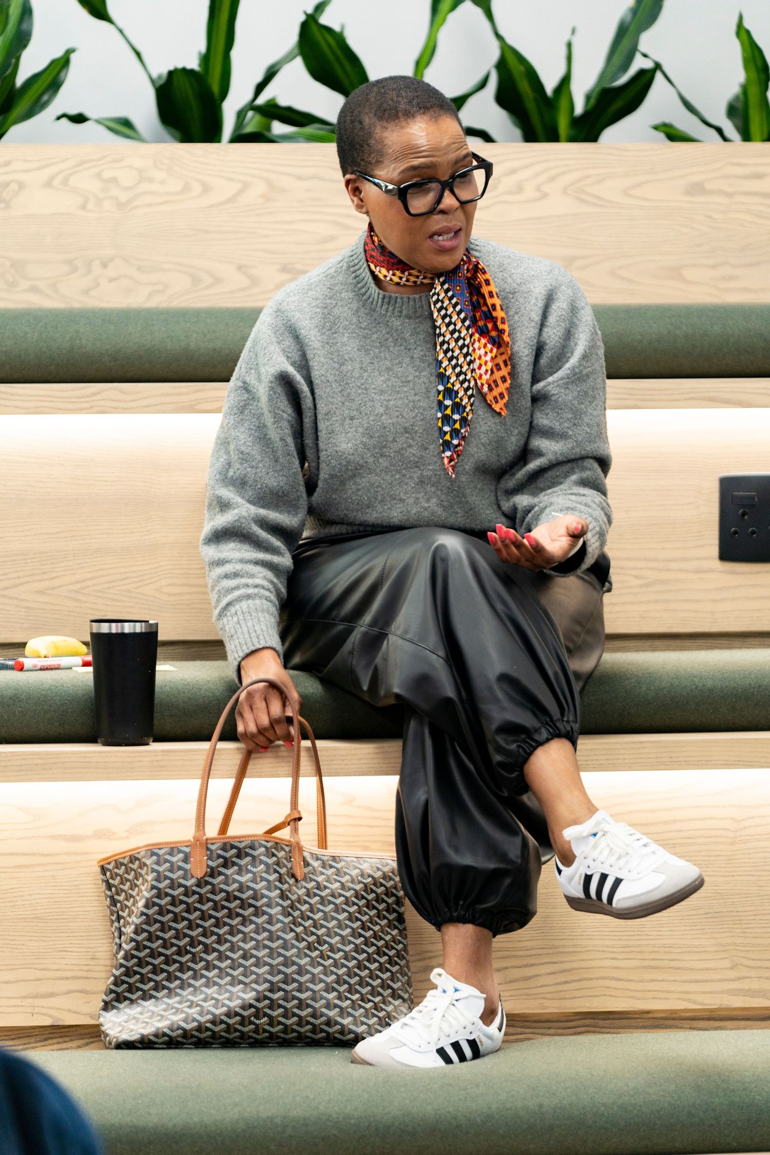 Stylish woman in gray sweater, black pants, and white Adidas sneakers sitting with a Goyard tote bag.