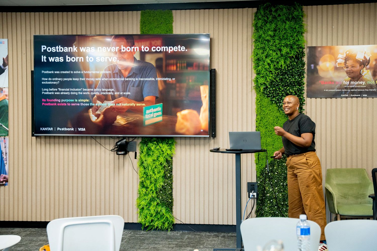 A presenter gestures toward a Postbank branded slide reading 'born to serve' during a corporate presentation in a modern office.