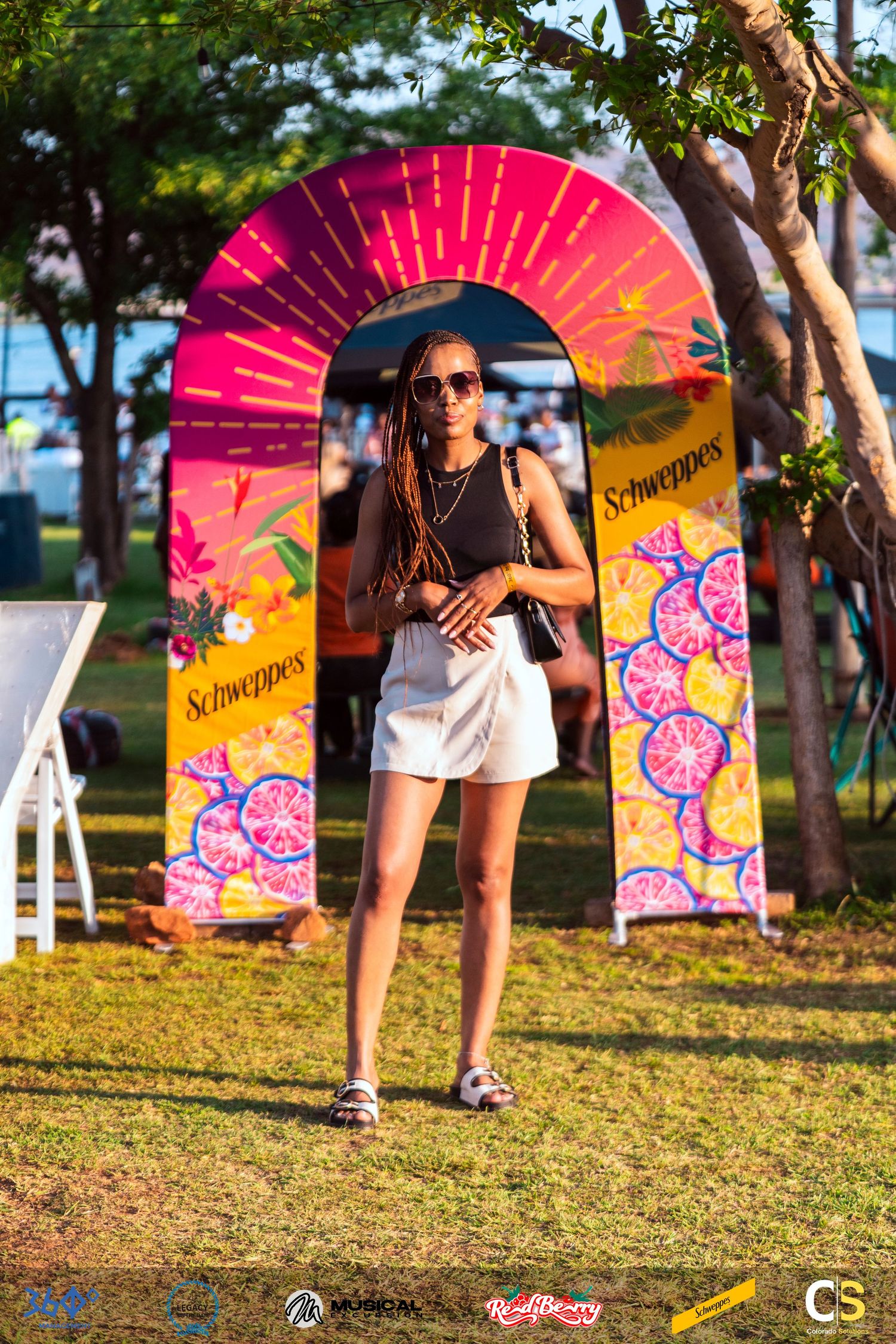 Woman in white shorts poses before a vibrant Schweppes branded arch display at an outdoor event.