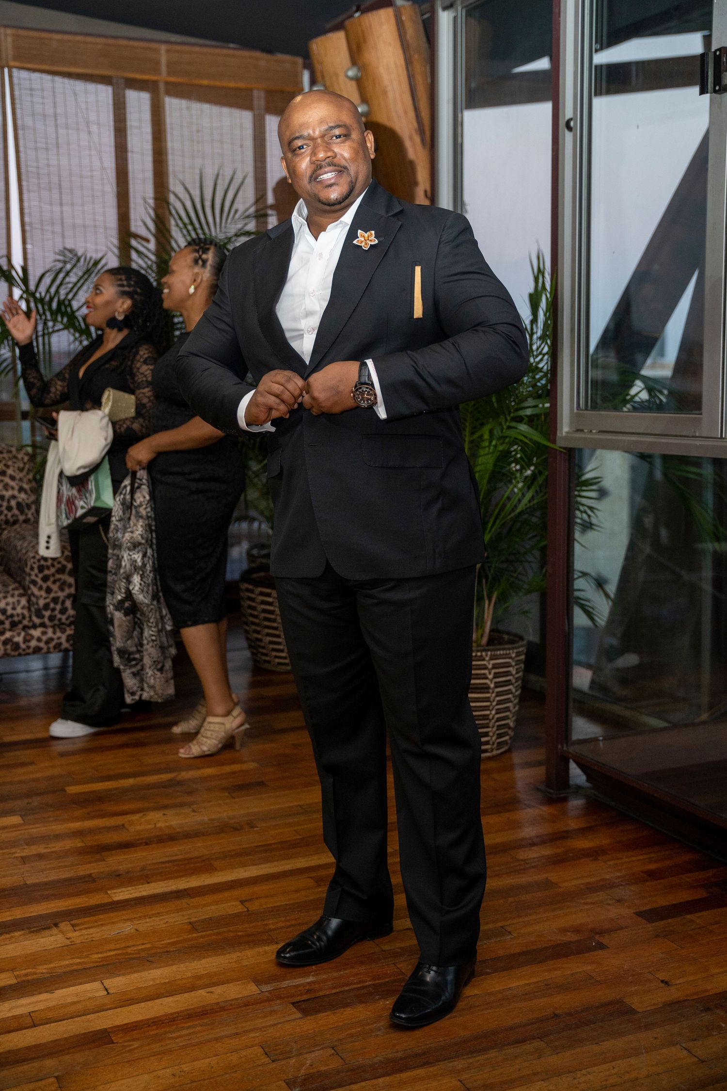 Smiling man in black suit with decorative pin posing at elegant indoor event.