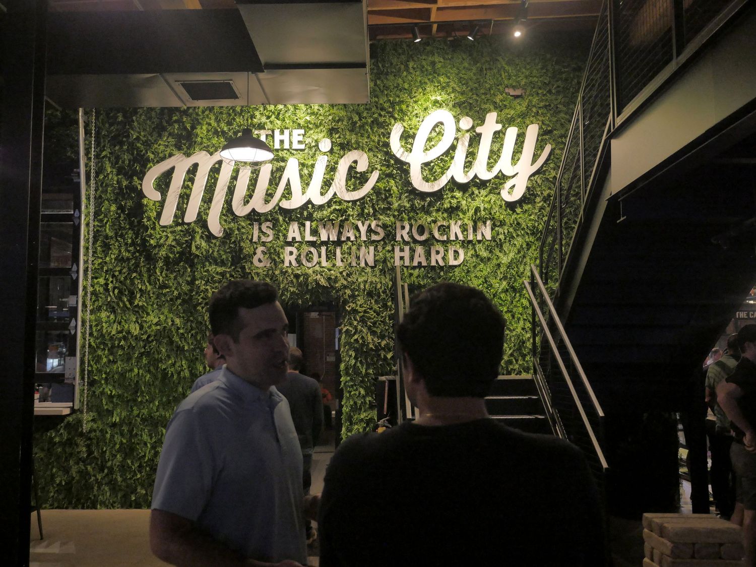 People gathered under illuminated Music City sign with green foliage wall.