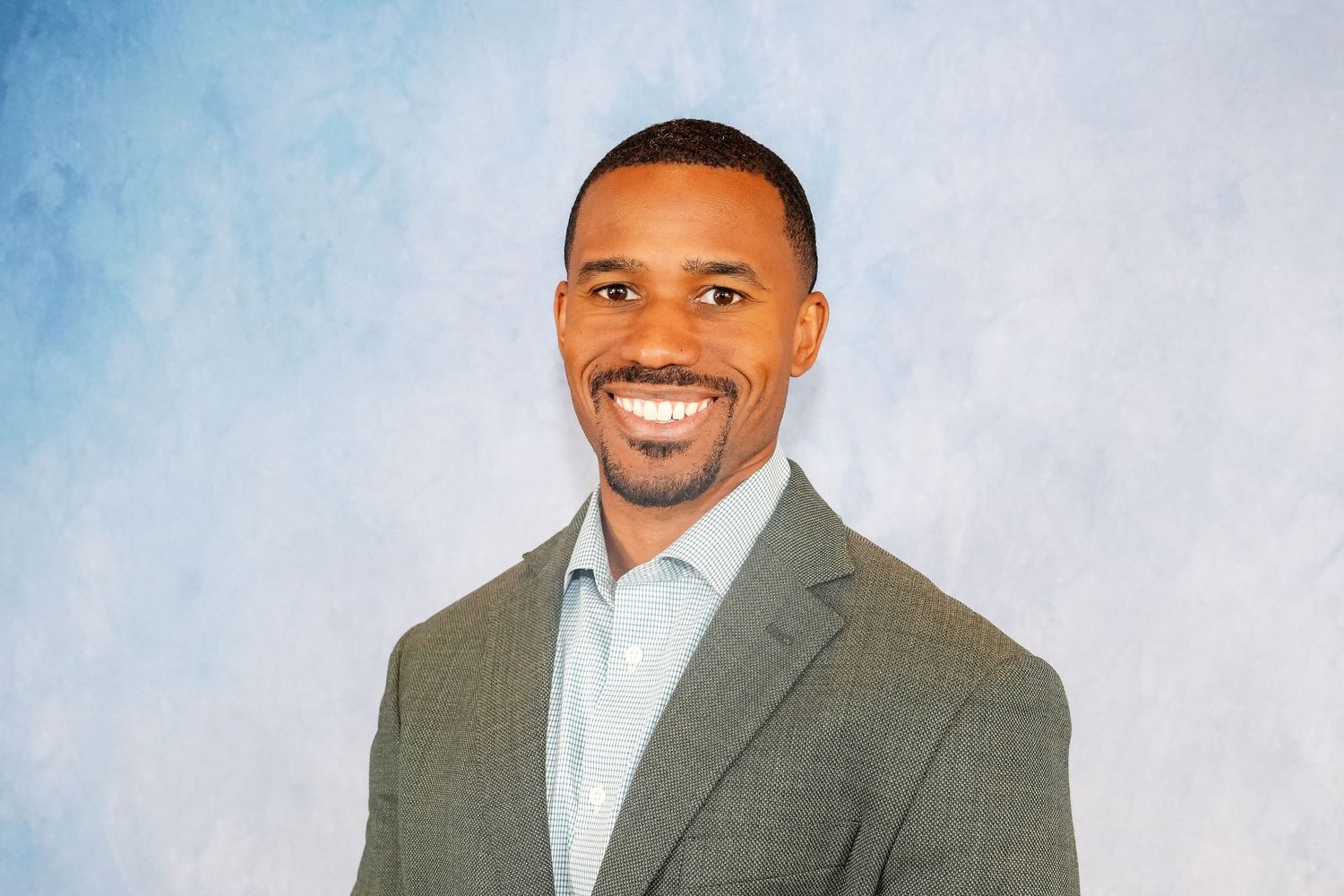 Professional headshot of person in grey suit jacket against blue background.