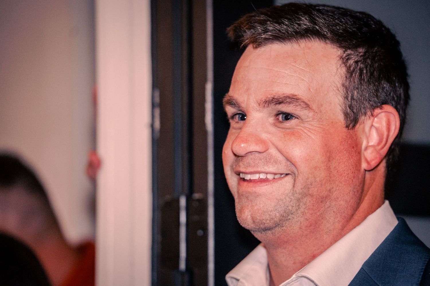 A person smiling warmly in a dark indoor setting.