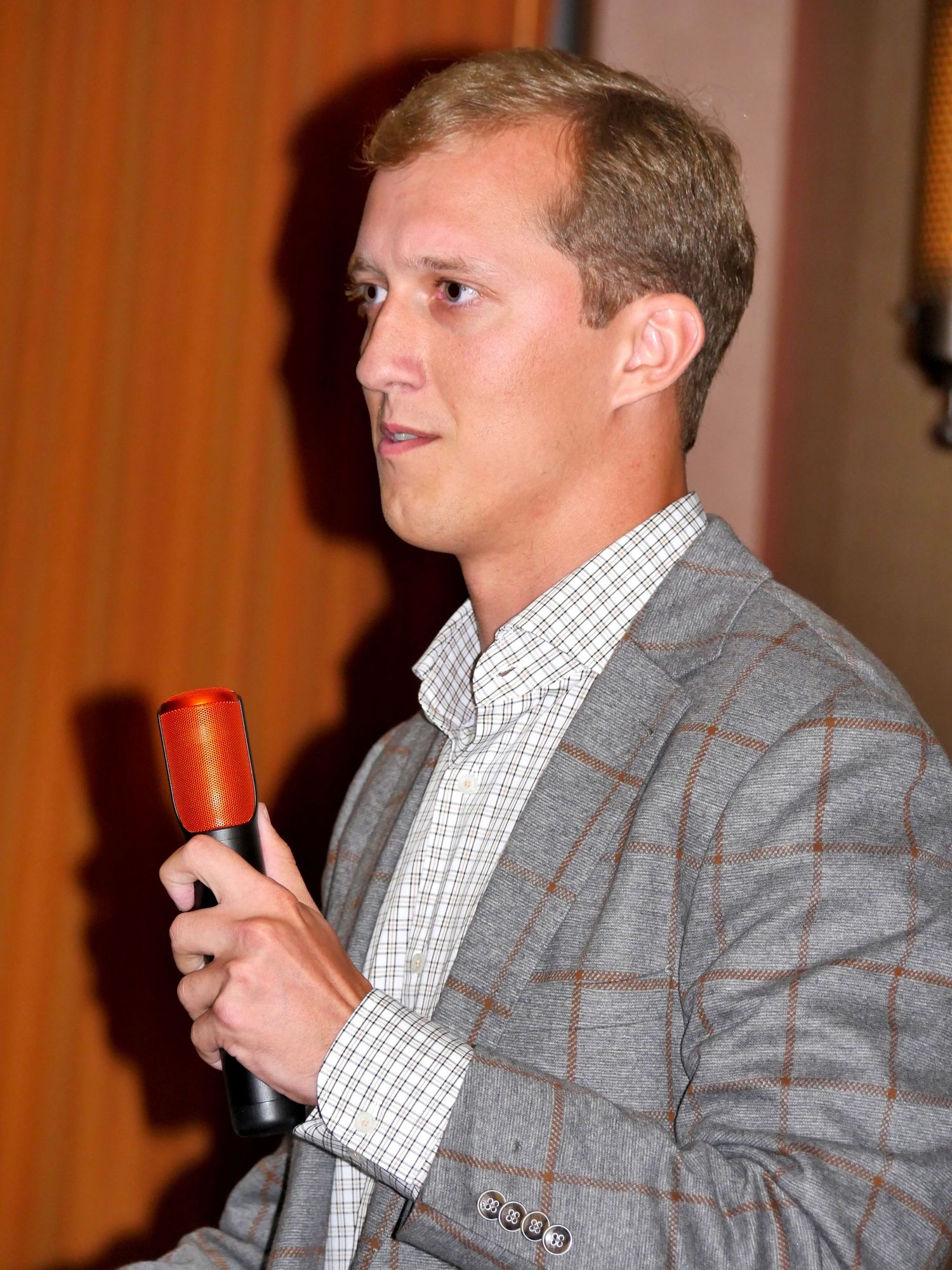 Person in checkered shirt and grey blazer speaking into microphone at event.