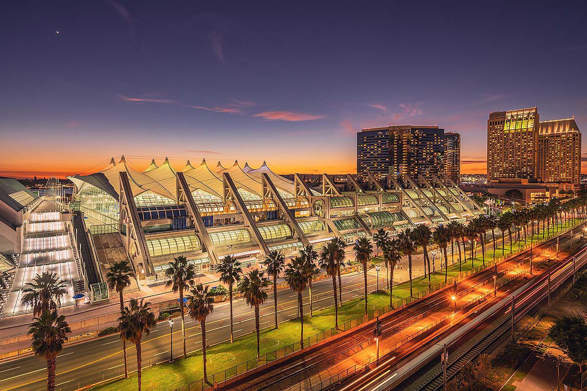 San Diego Convention Center illuminated at sunset with palm trees and city buildings along Harbor Drive.
