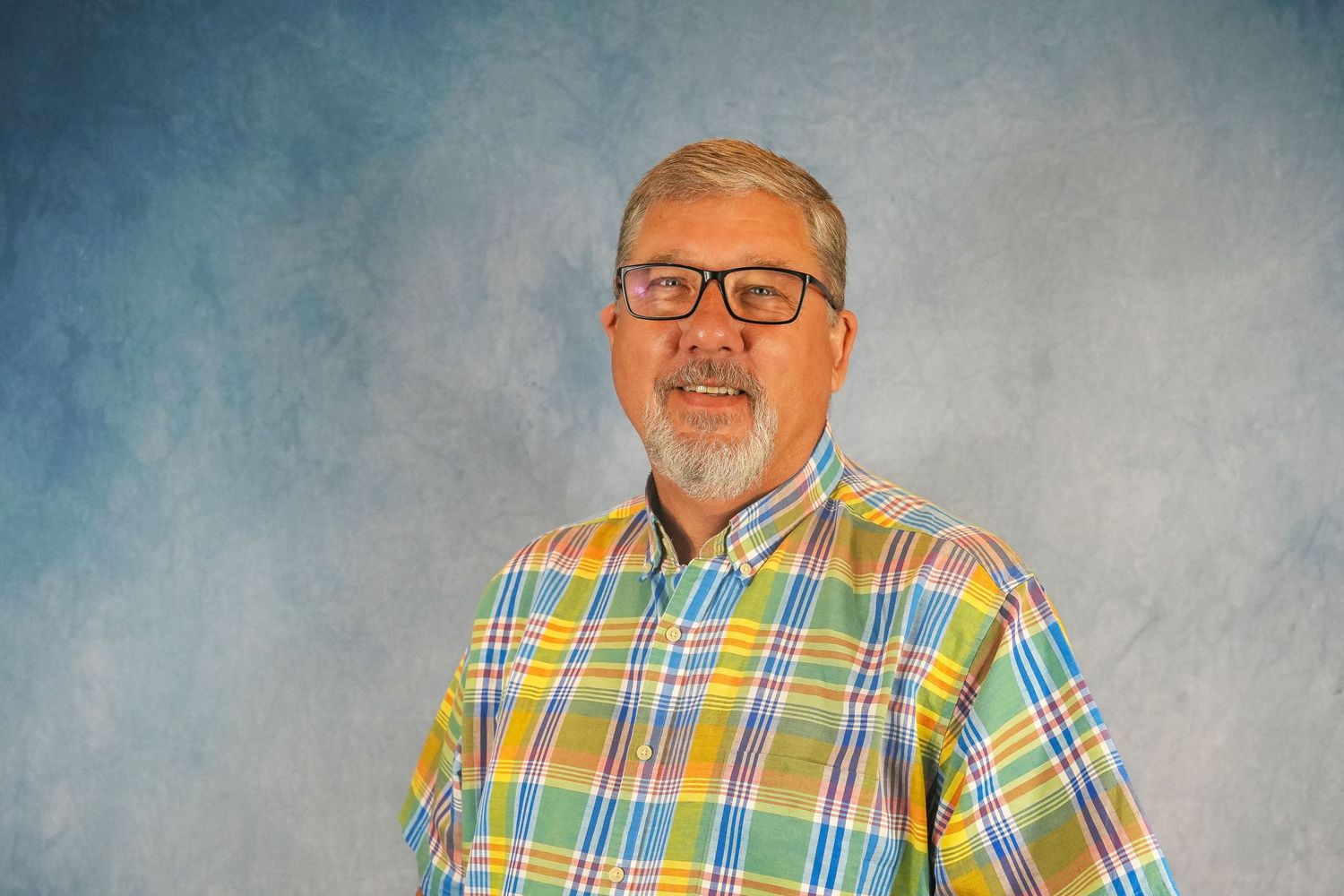 Professional headshot of person in colorful plaid shirt against blue background.