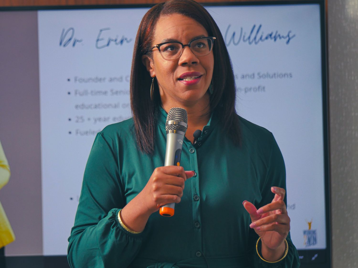 Business presenter speaking into microphone with presentation slides in background.