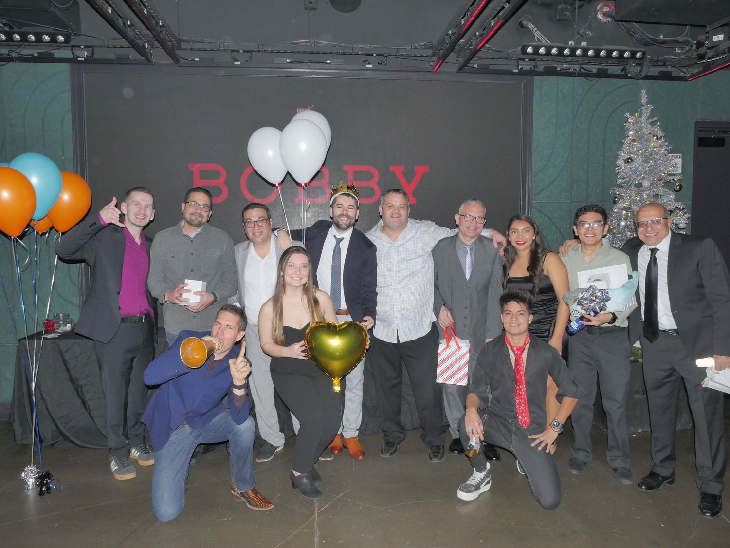 Group photo at a party with colorful balloons and a white Christmas tree in the background.