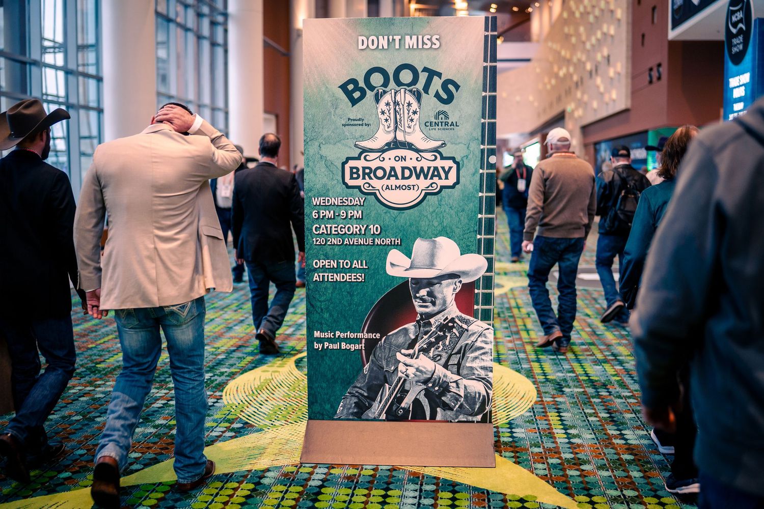 People walking through a convention center hallway with blue and green patterned carpet and branded signage.