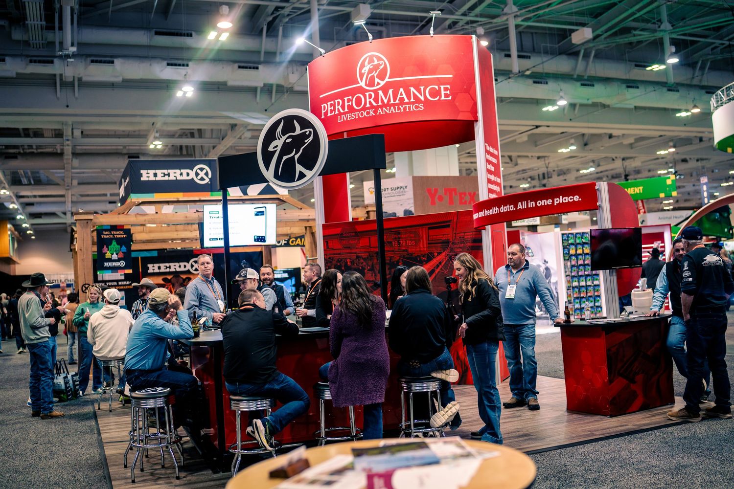 Trade show exhibition hall with red and black branded booths and attendees walking through the convention space.