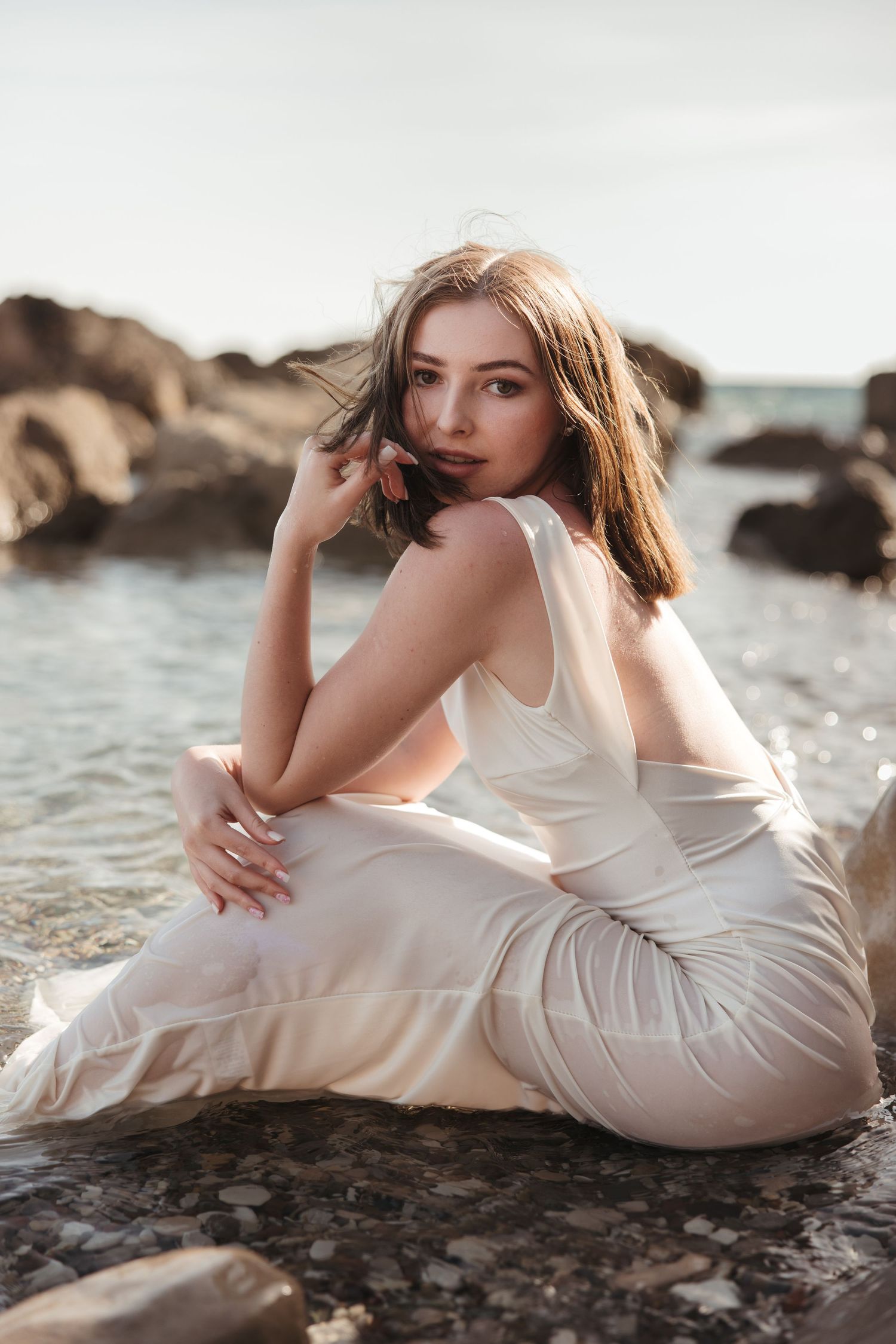 Woman in a white dress sitting on rocky coastline during a sunset photoshoot in Cyprus