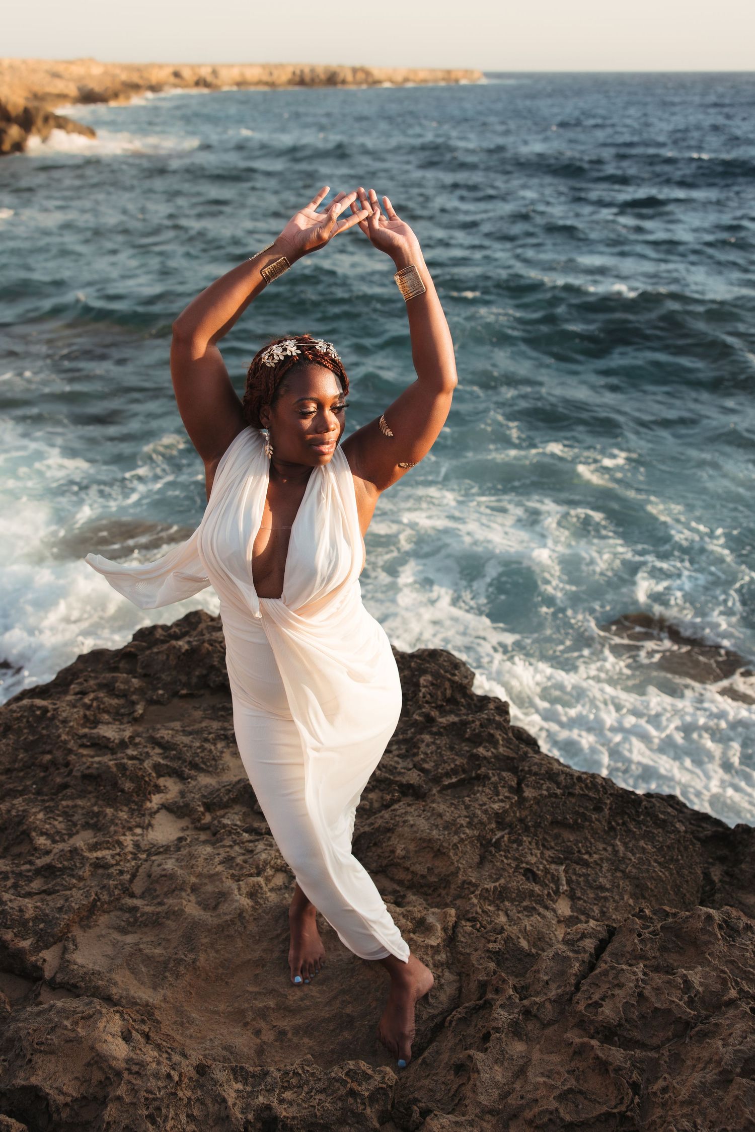 Woman in a white dress standing on coastal rocks with the Mediterranean Sea behind her during a Cyprus beach photoshoot