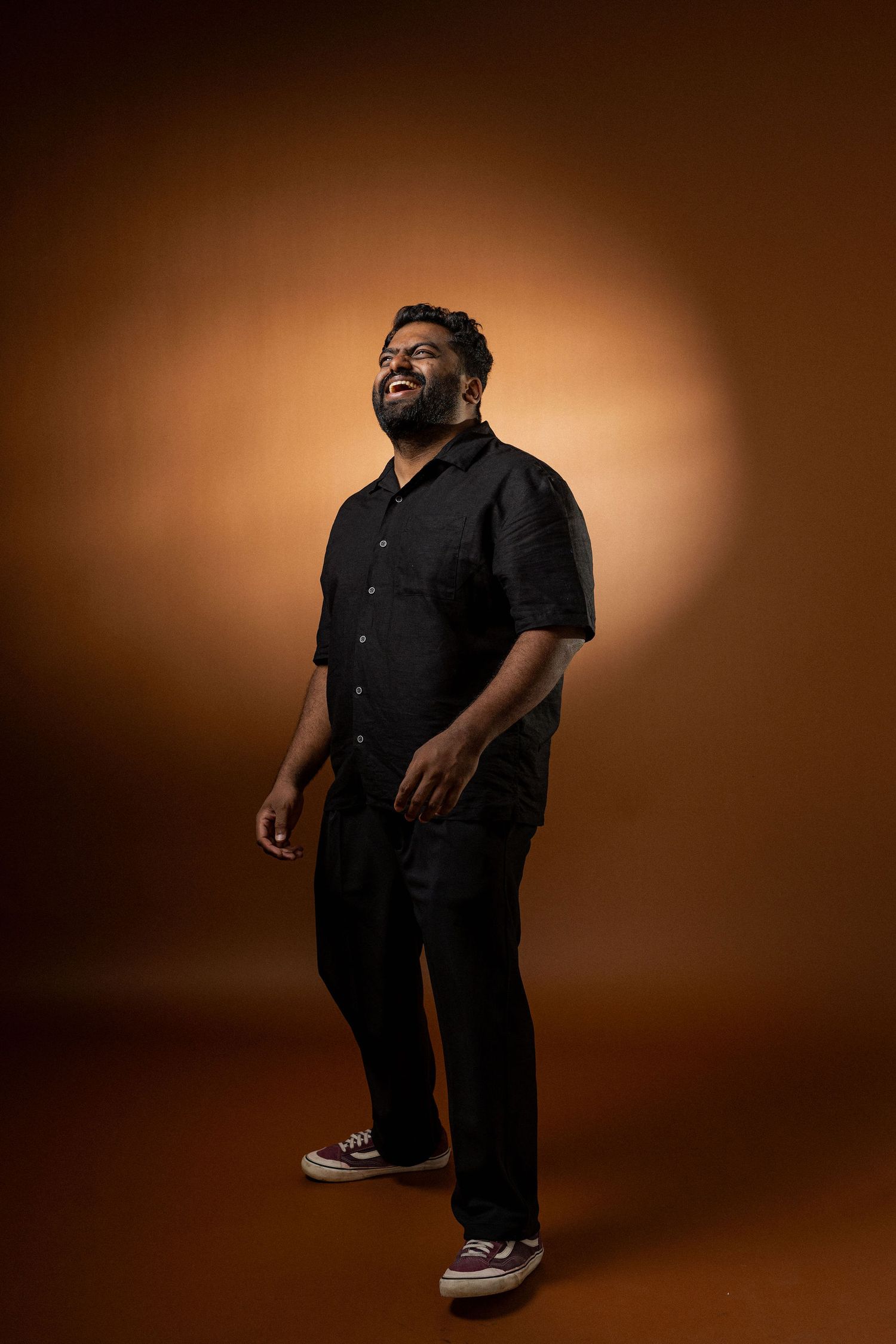 A person wearing all black clothing and sneakers poses against a warm brown studio backdrop with dramatic lighting.