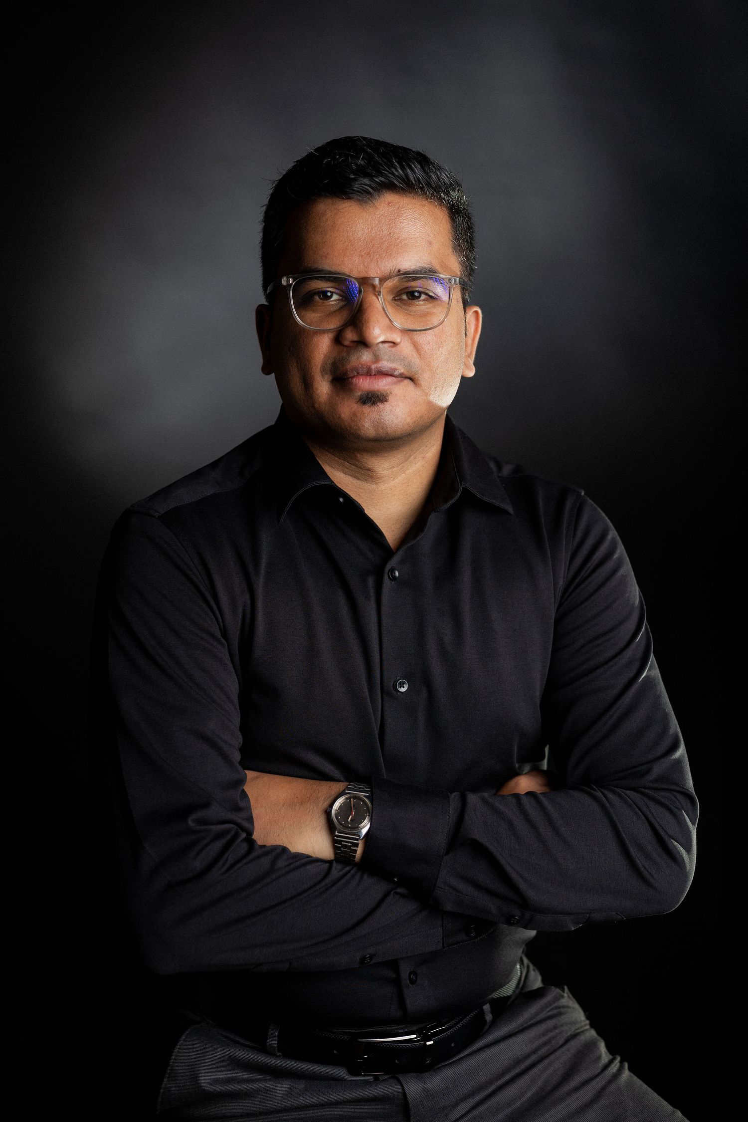 Professional studio portrait of person in black button-up shirt against dark background in dramatic lighting.