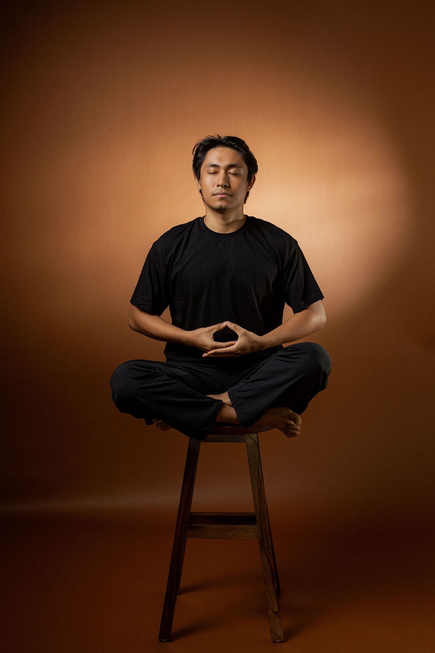 A person in a black t-shirt sits cross-legged in meditation pose on a wooden stool against a brown backdrop.