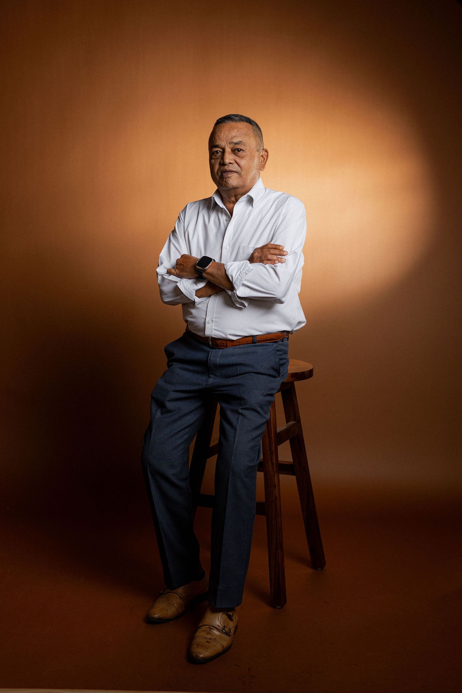 Studio portrait of person in white shirt and dark pants sitting on wooden stool against warm brown backdrop.
