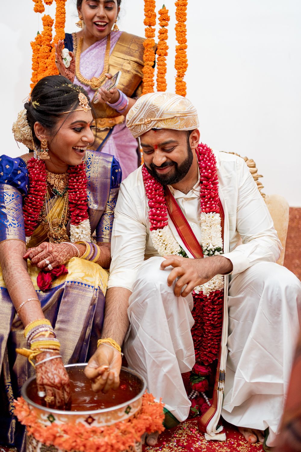A couple participates in a traditional Indian wedding ritual with orange and red flower garlands and ceremonial items.