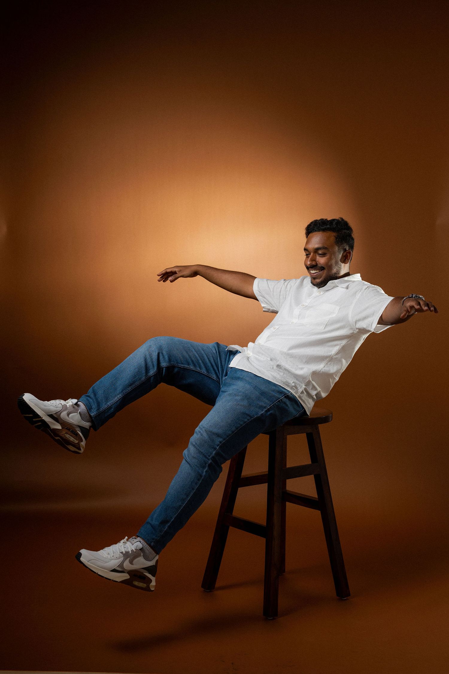 Person in white t-shirt and jeans reclines dramatically on dark wooden stool against brown backdrop in sequence of poses.