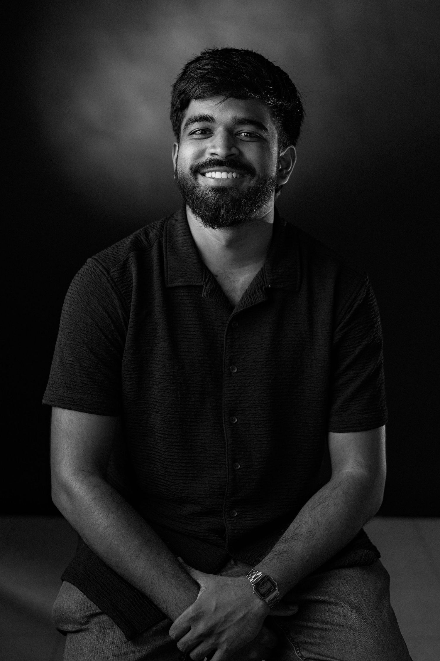 A black and white portrait of someone in a dark polo shirt smiling warmly against a shadowy background.