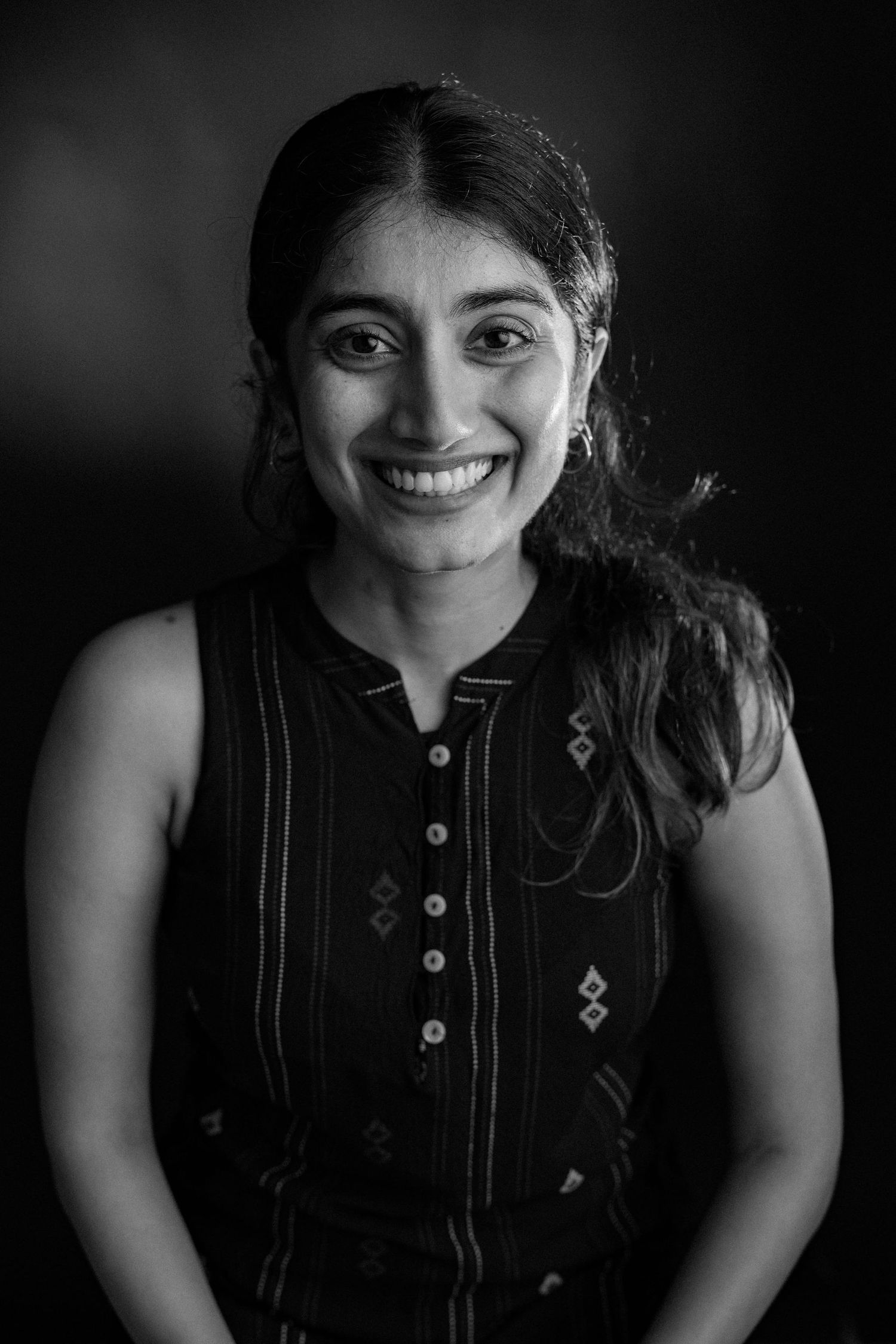 Black and white portrait of someone wearing a sleeveless button-up top with embellished details, smiling warmly at camera.