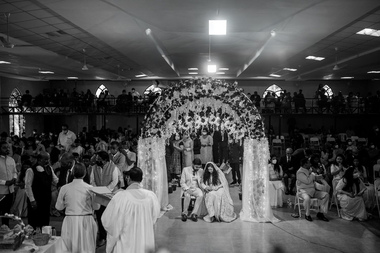 A black and white wedding ceremony photo showing a decorated arch and crowd gathered in a large indoor venue.