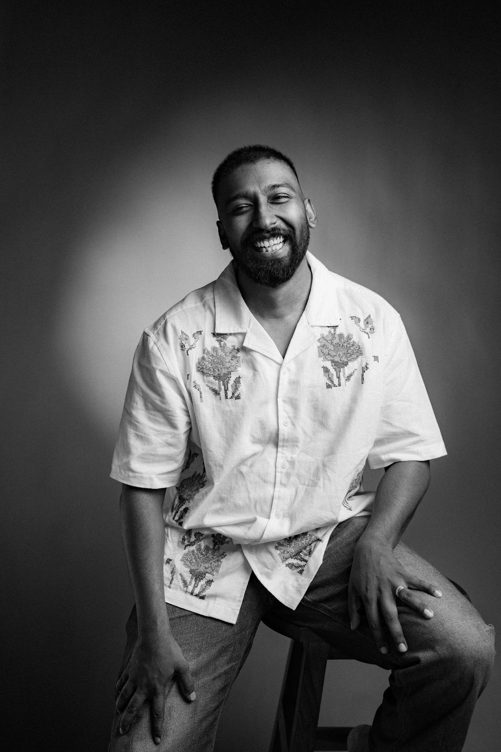 Black and white portrait of a smiling bearded man in a floral shirt, seated against a gradient background.