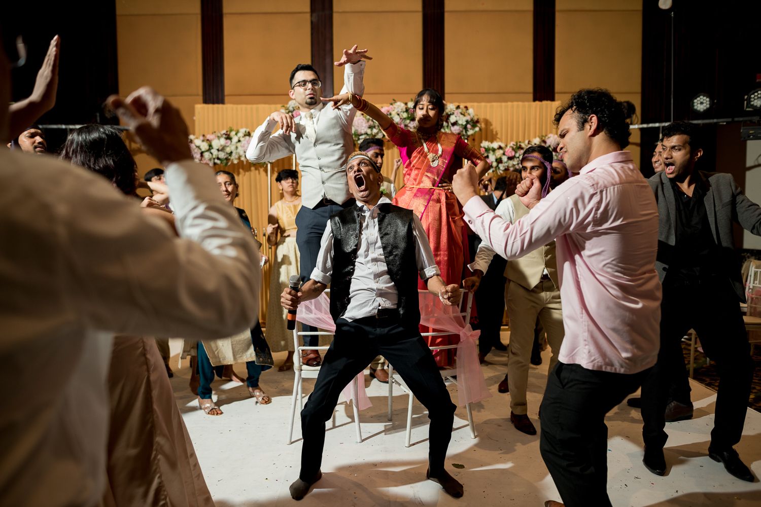 Wedding guests dance enthusiastically on a dance floor during a reception celebration in an indoor venue.