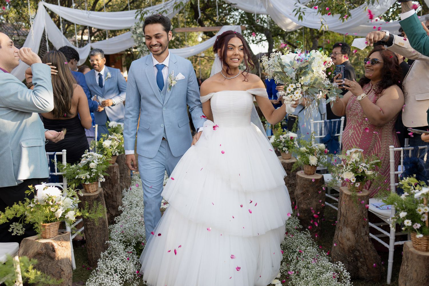 Wedding guests toss pink rose petals as newlyweds walk down garden path at outdoor ceremony celebration.