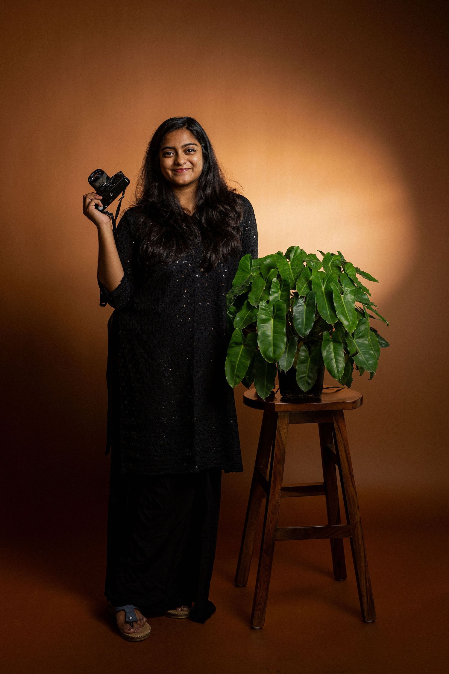 A photographer stands in a dark dress next to a potted plant on a wooden stool against a warm brown background.