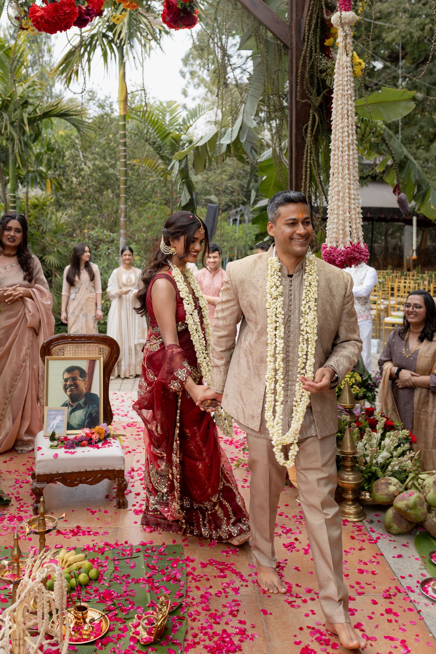 Traditional Indian wedding ceremony with rose petals scattered on aisle and red floral decorations hanging from above.