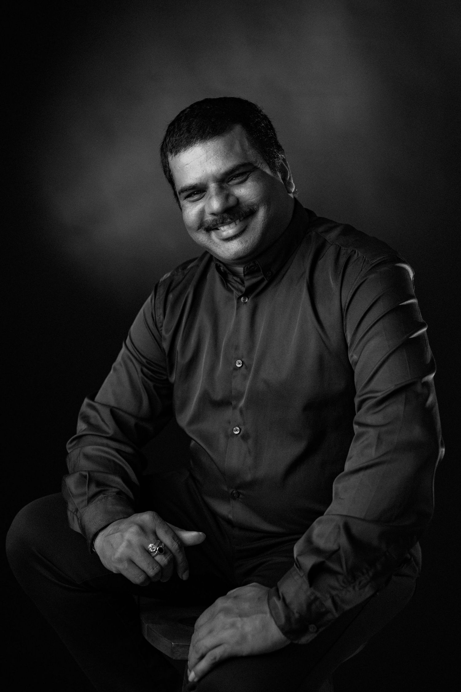 Black and white portrait series showing a person in dark clothing smiling and looking down while sitting against dark background.