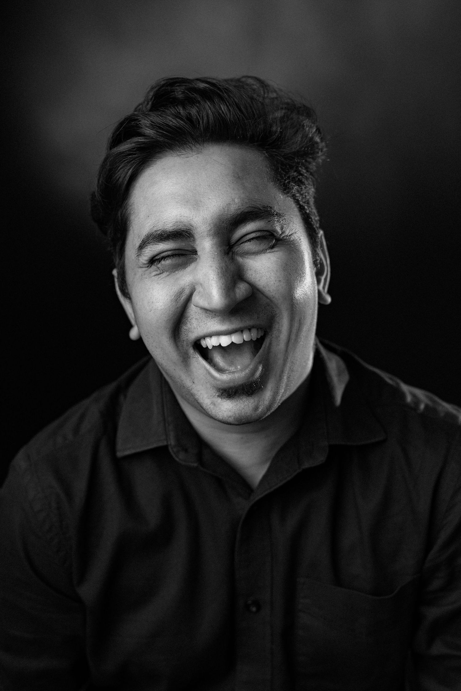 A black and white portrait of someone laughing joyfully with their eyes closed against a dark background.
