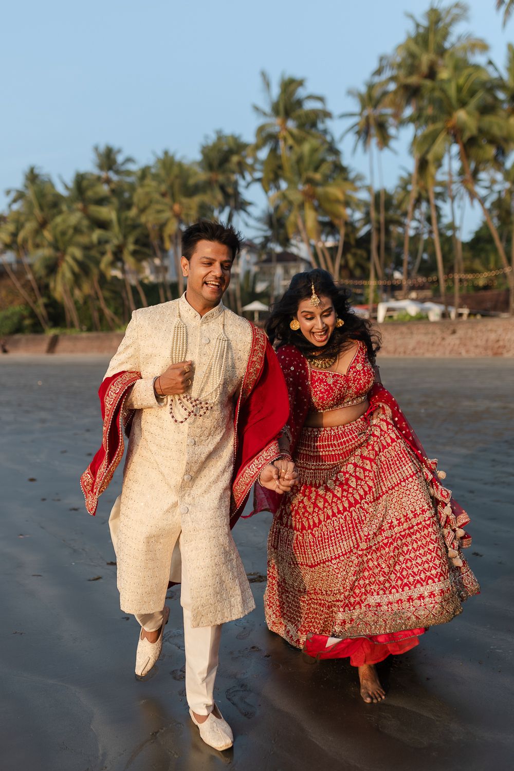 A couple in traditional Indian attire walking joyfully on a beach with palm trees in the background.