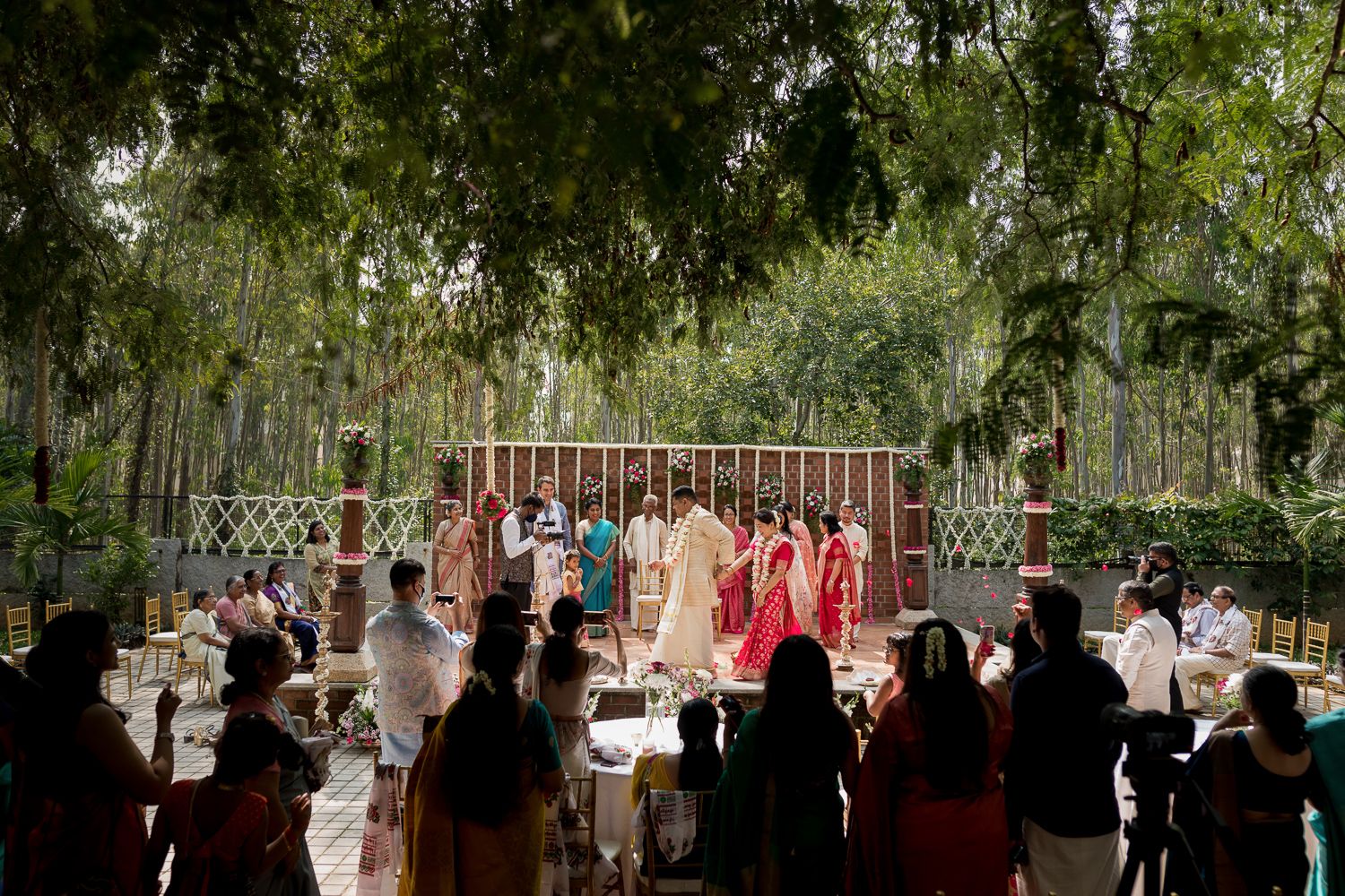 Wedding guests gather outdoors under tall trees for a ceremony with white chairs and decorative elements.