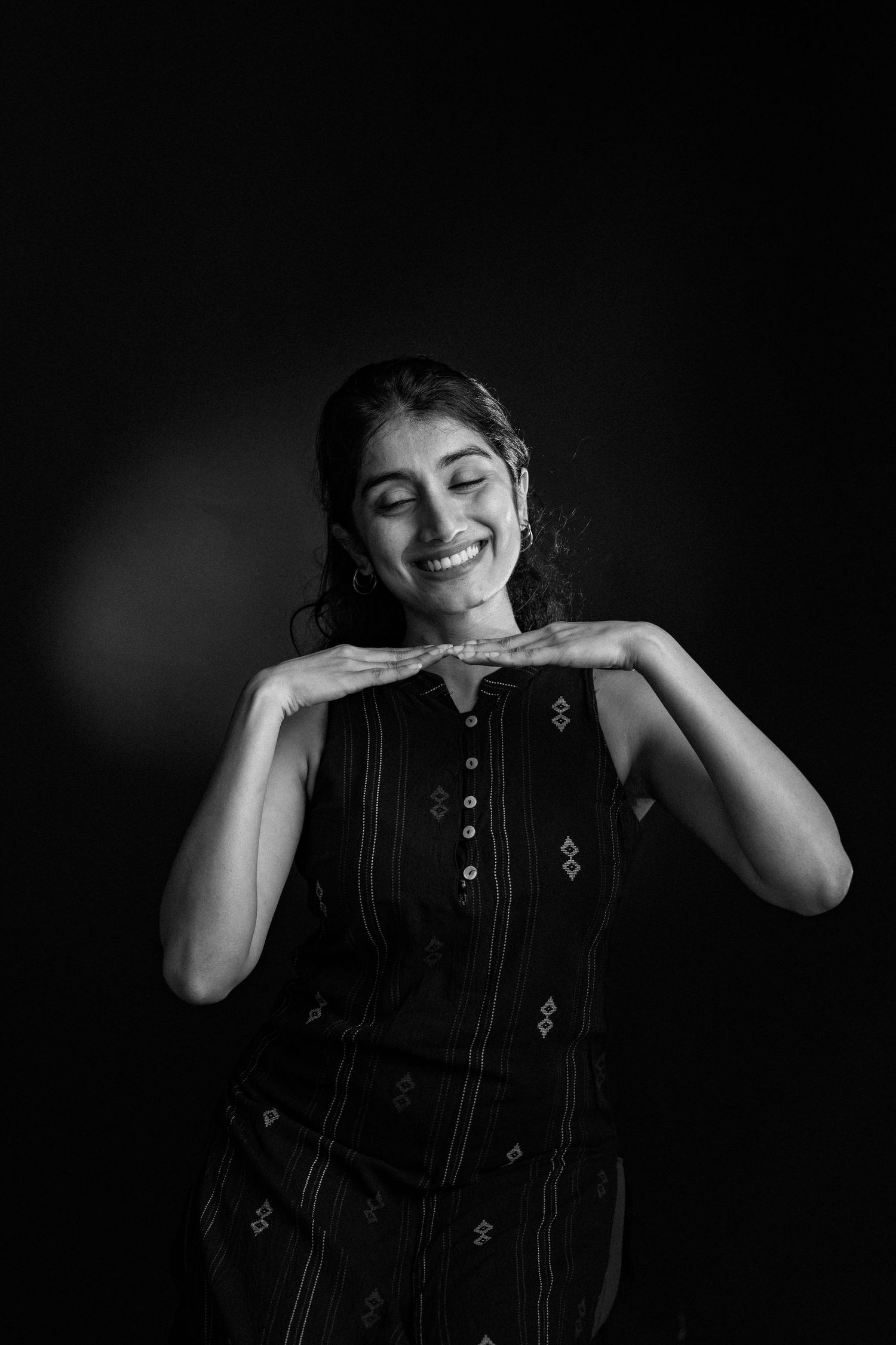 Black and white portrait of someone in a patterned dress smiling and resting their chin on their hands against dark background.