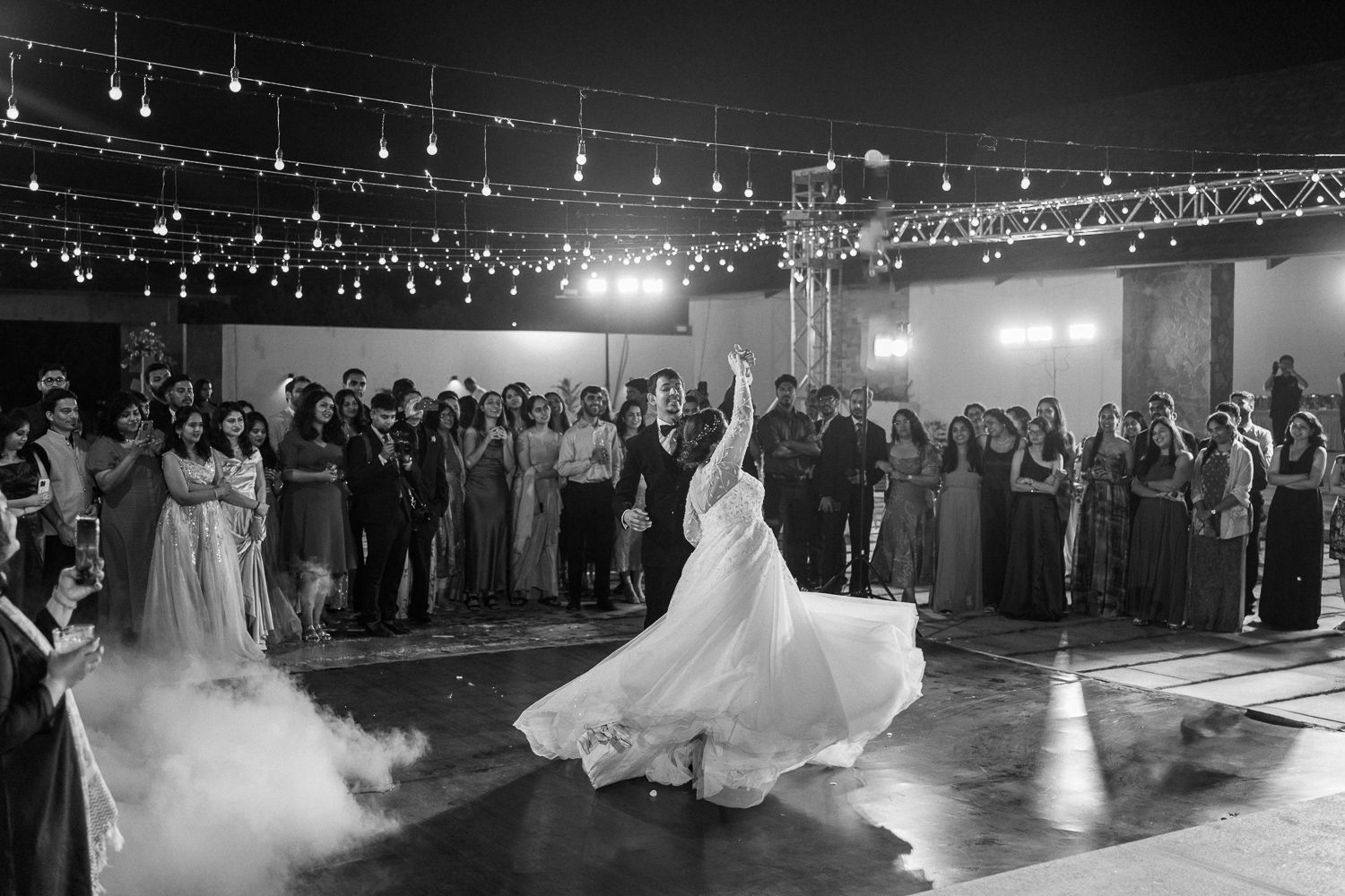 Wedding couple dances under twinkling lights at their reception while guests watch from the dance floor.