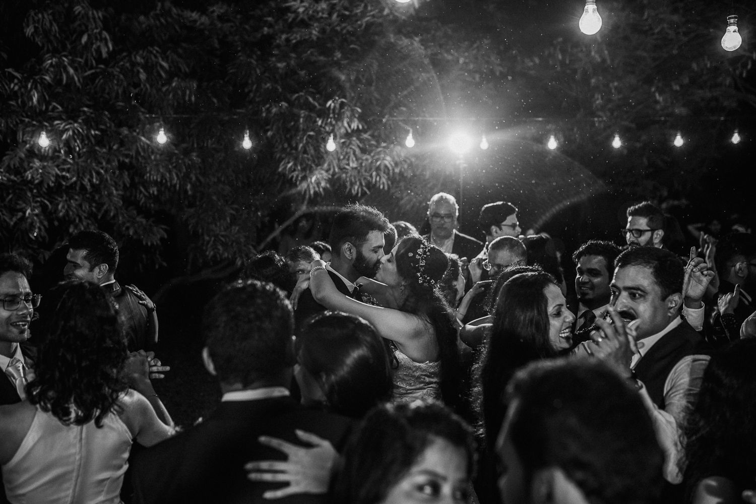 A romantic dance moment captured in black and white under glowing string lights at an outdoor evening celebration.