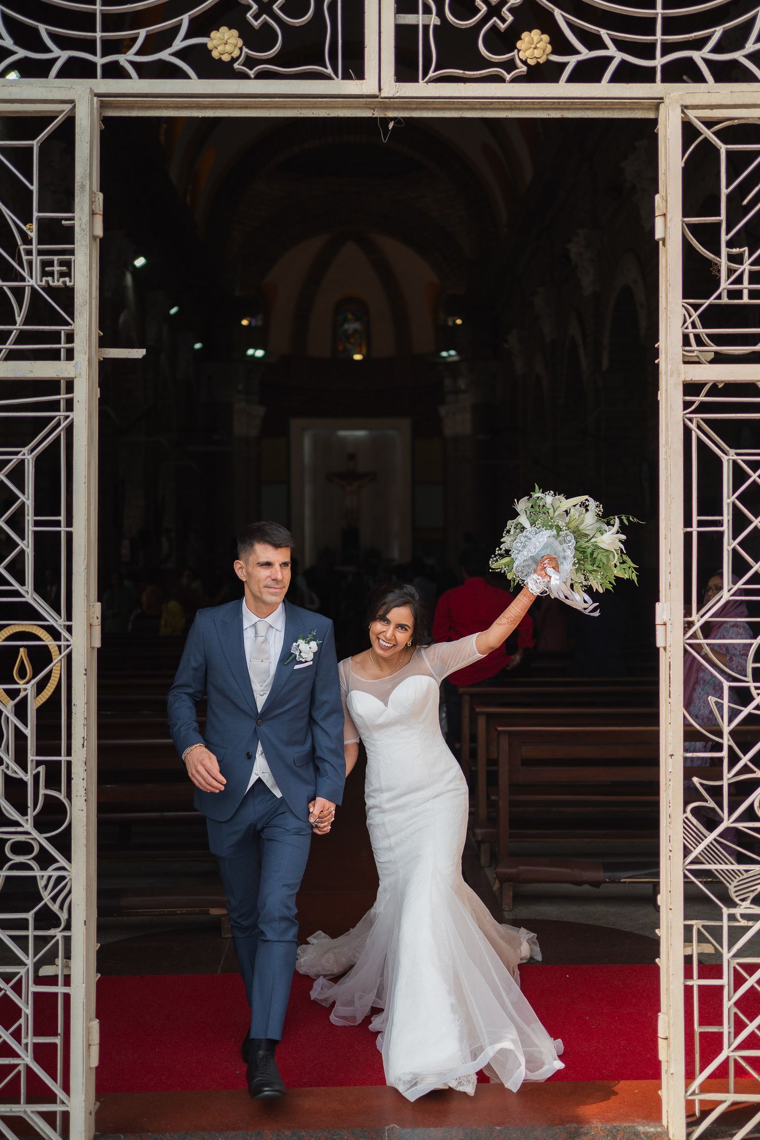 A joyful wedding couple exits their ceremony venue walking down a red carpet under decorative white archways.